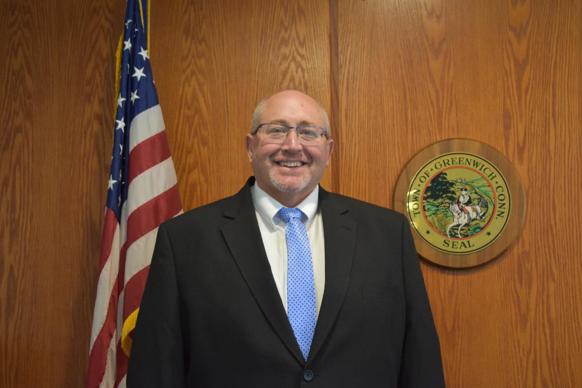 James Michel, newly appointed Commissioner of the Department of Public Works for the Town of Greenwich, stands in front of the town seal and the American flag, wearing a suit and blue tie, smiling confidently.