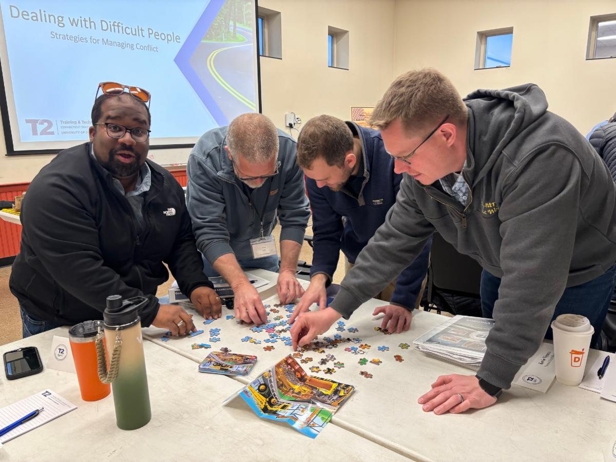 four people working together on a puzzle during a dealing with difficult people workshop with a presentation screen in the background displaying strategies for managing conflict