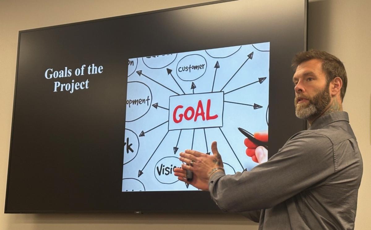 man giving presentation pointing to screen that says goals of the project with diagram showing arrows leading to a center box labeled goal