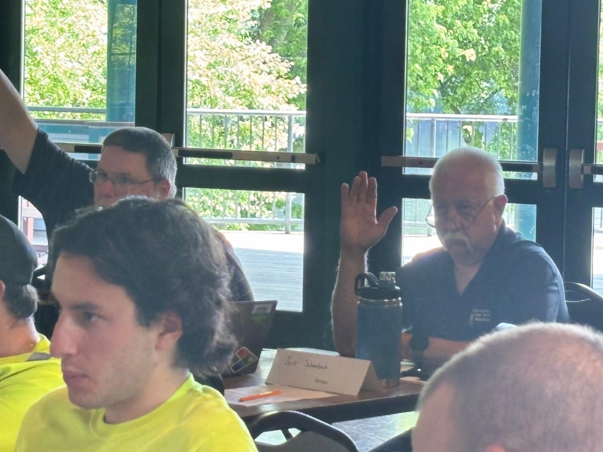 man sitting at a table with hand raised during a workshop session with other participants listening and large windows showing trees in the background