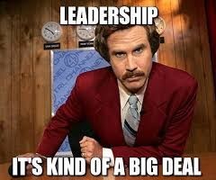 man in a burgundy suit leaning on a desk with text that reads leadership its kind of a big deal