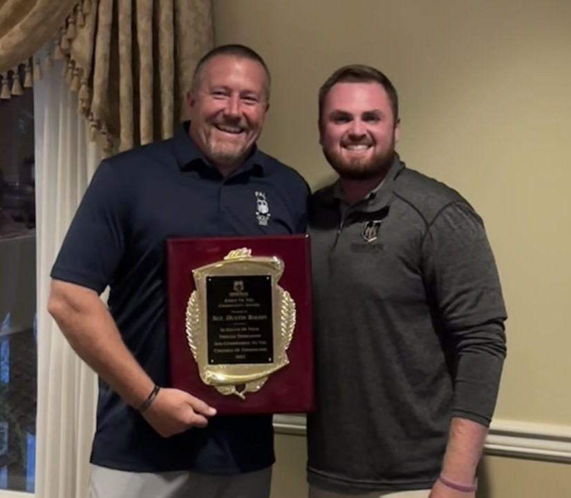 two men standing indoors smiling as one holds a large plaque award with gold detailing against a cream-colored wall