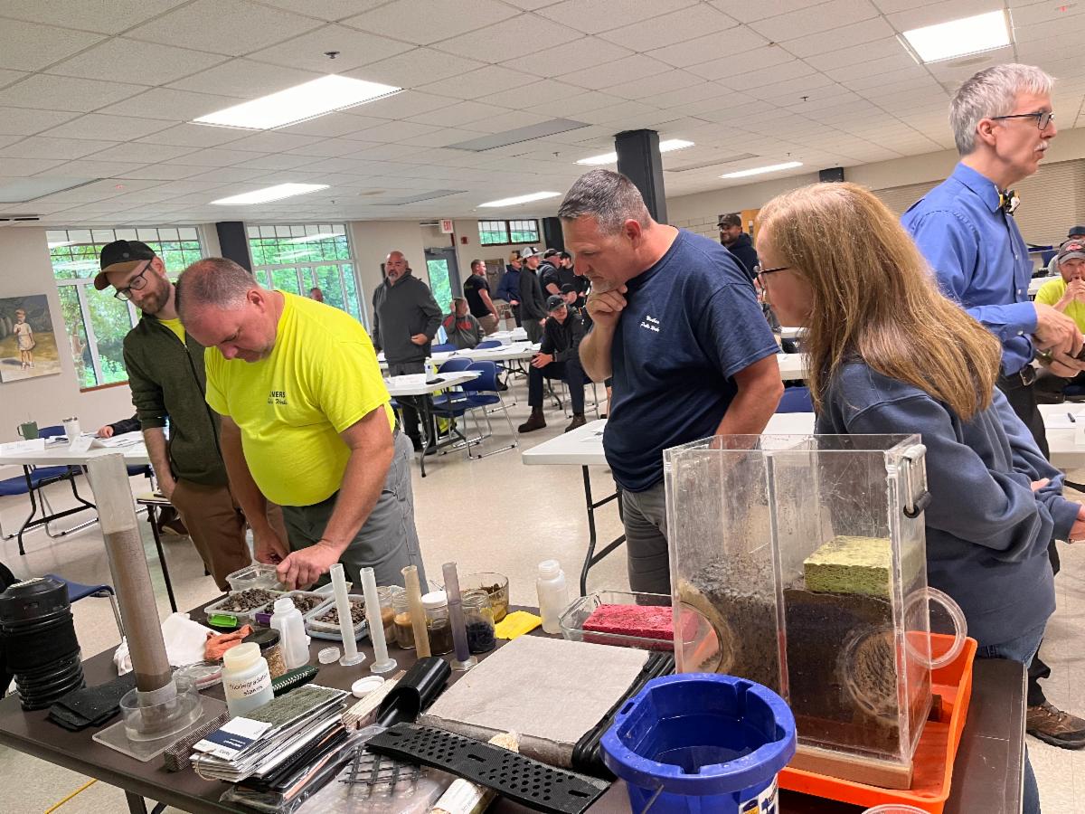 Participants sit around a table in a large room actively engaged in a hands on training exercise using a tabletop drainage layout.