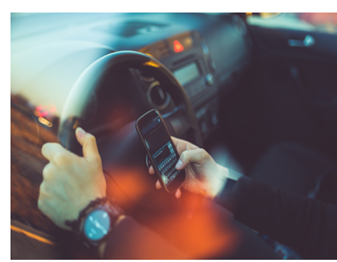 A person driving a car while holding a smartphone in one hand and steering with the other, suggesting distracted driving. The interior of the car is visible, with a watch on the driver's wrist and soft light reflections on the image.