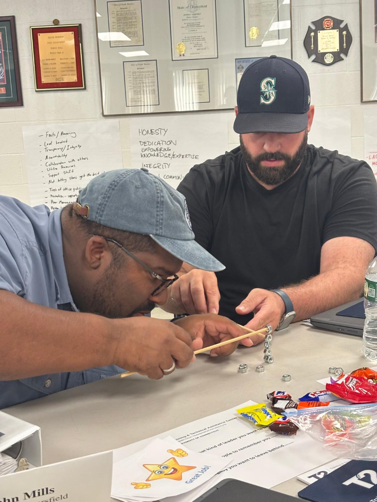 two men work together on a hands on activity at a table one man is focused on balancing metal nuts with a wooden stick while the other watches and assists papers candy and water bottles are on the table and posters with words like honesty dedication and in