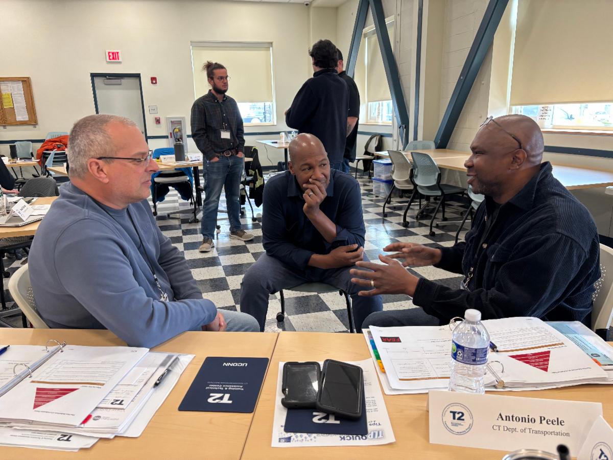 Three men sit at a classroom table engaged in a focused discussion during a workshop. Printed materials, notebooks, and a nameplate are spread across the table. In the background, other participants are standing and talking in a well-lit room with checkered flooring and large windows.