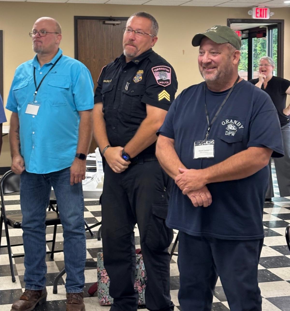 three men standing and smiling in a classroom setting one wearing a blue shirt one in a police uniform and one in a granby dpw shirt with people and chairs in the background