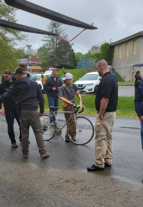 People gathered around a bicyclist during a roadway safety demonstration, with participants observing and discussing a simulated bicycle crash scene.