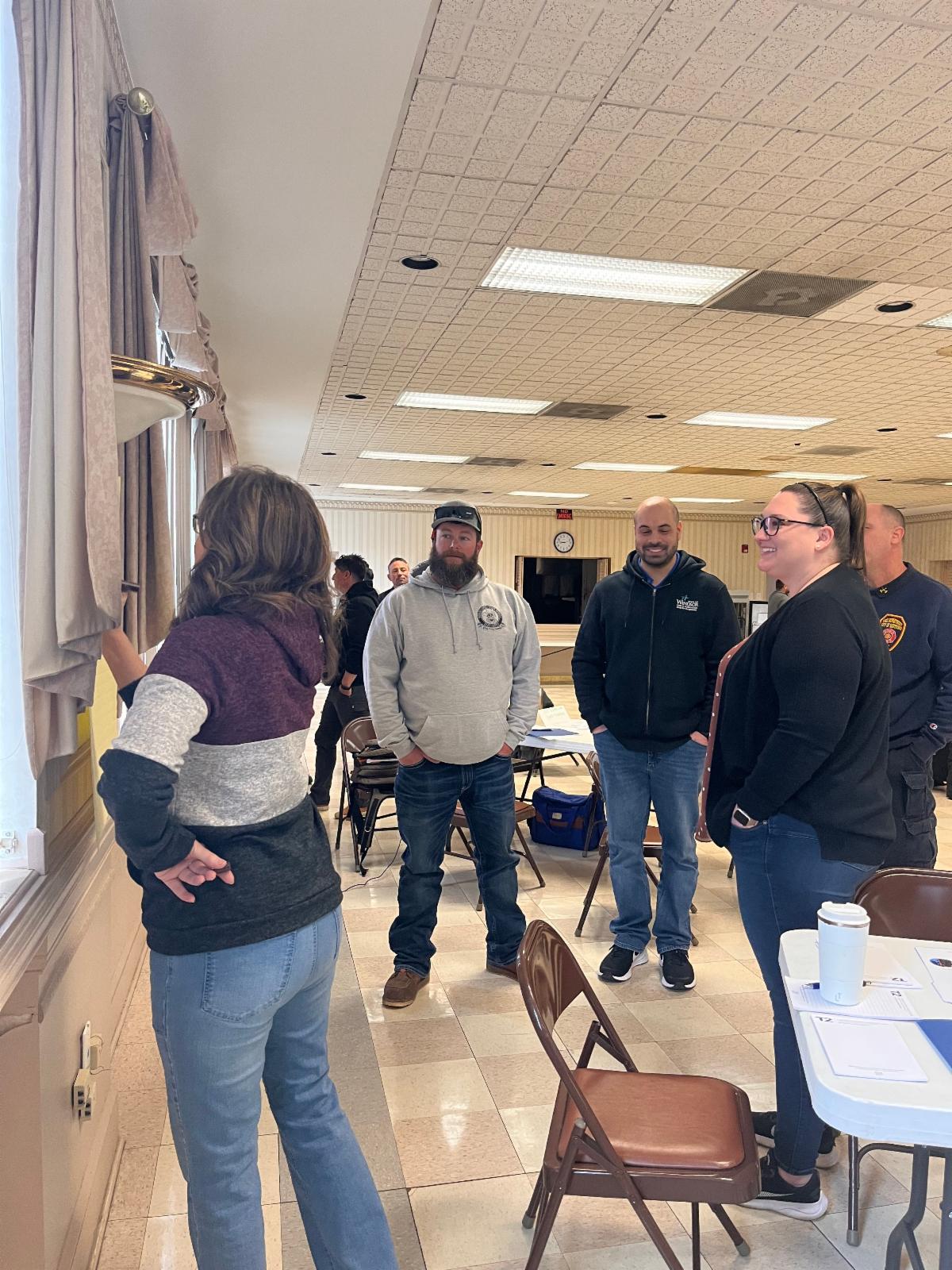 A group of adults stands inside a community room, engaged in discussion while looking at something posted on a wall.