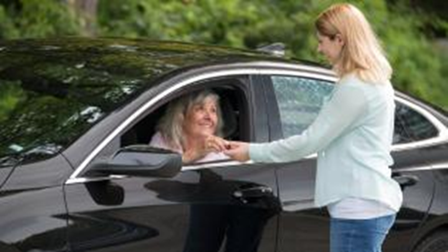 woman standing outside handing an item to another woman seated in a car smiling at each other in a parking area with trees in the background