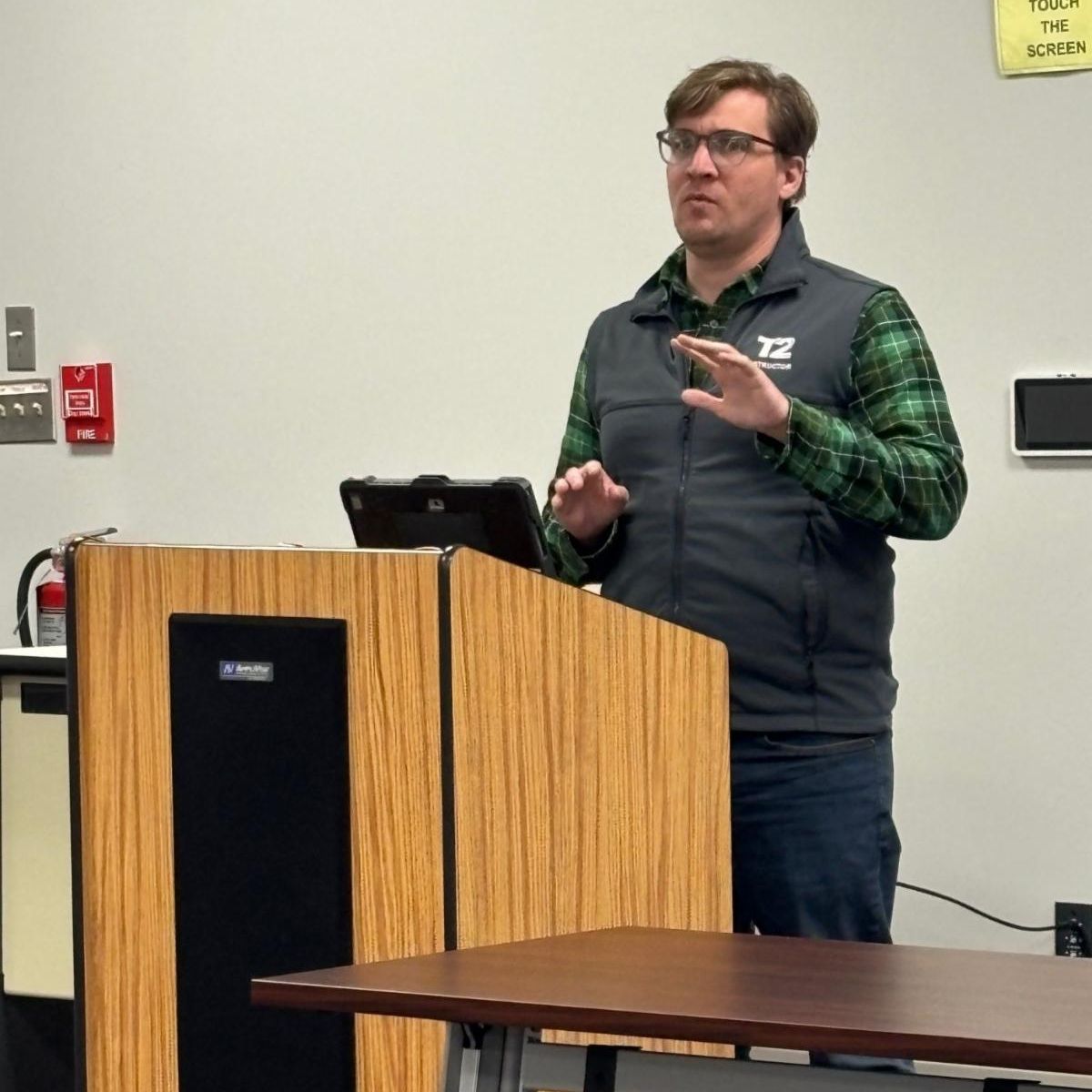man wearing glasses and a t2 instructor vest speaking at a wooden podium in a classroom setting with a sign on the wall that says do not touch the screen