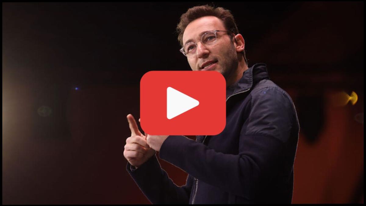 Man wearing glasses and a dark jacket speaking on stage with one hand raised and pointing large red play button icon over image suggesting a video thumbnail