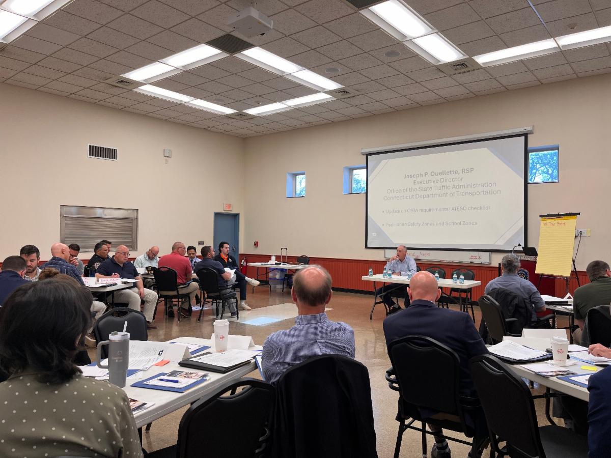 Participants sit at round tables in a large, well-lit conference room, attentively listening to a presentation projected on a screen at the front of the room. Workshop materials and nameplates are visible on the tables, indicating a formal training or professional development session.