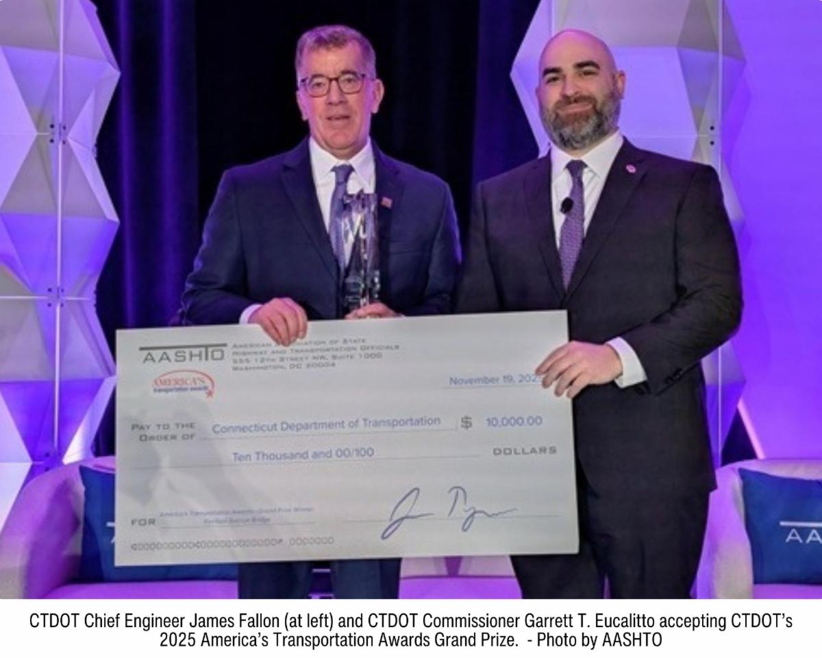 two men in suits standing on stage holding a large ceremonial check with CTDOT identified as the recipient against a backdrop of geometric panels and purple lighting