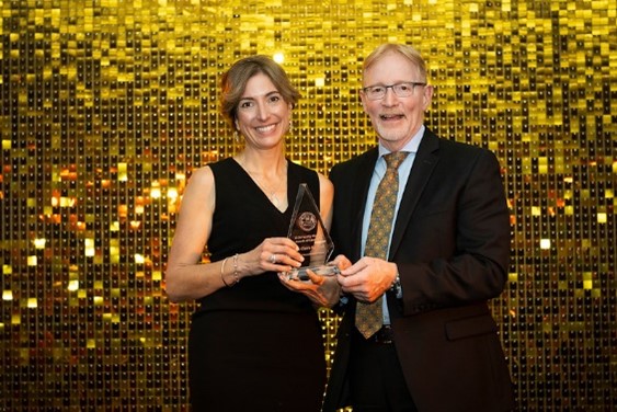 Dr Claire Kendall standing in front of a gold disco ball photo background holding her award and standing beside the Dean Dr Bernard Jasmin