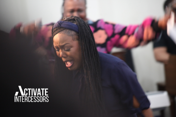 Dianna Hobbs with long braids and a headband dances in the sanctuary, her face lifted in passionate worship, mouth open in praise. She wears a navy top, and behind her, others join in the move of God, one in a colorful floral dress. The text “ACTIVATE INTERCESSORS” is bolded in white over a black background in the bottom left corner, capturing the event’s power.