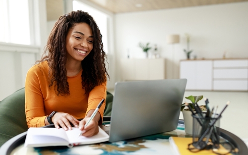 Smiling young African American woman sitting at desk working on laptop taking notes in notebook_ happy millennial female studying online_ watching webinar using computer and writing check list