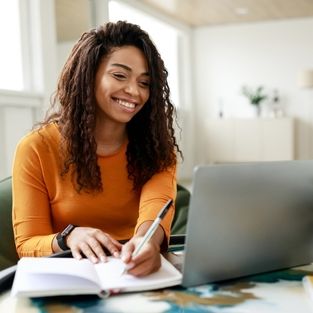 Smiling young African American woman sitting at desk working on laptop taking notes in notebook_ happy millennial female studying online_ watching webinar using computer and writing check list