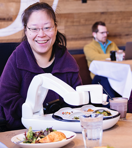 A smiling woman positioned in a wheelchair in front of the Obi Feeder full of food in a restaurant.