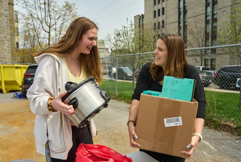Two students smile at each other as they hold boxes of items