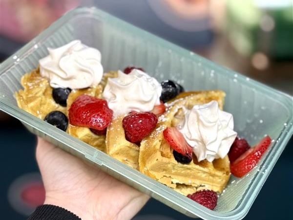 A hand holds a container with waffles topped with cream and berries