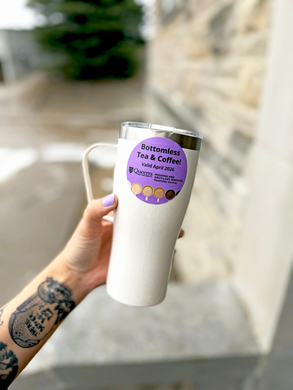 A tattooed arm holds up a reusable mug with a purple Bottomless Tea and Coffee sticker valid for April 2026