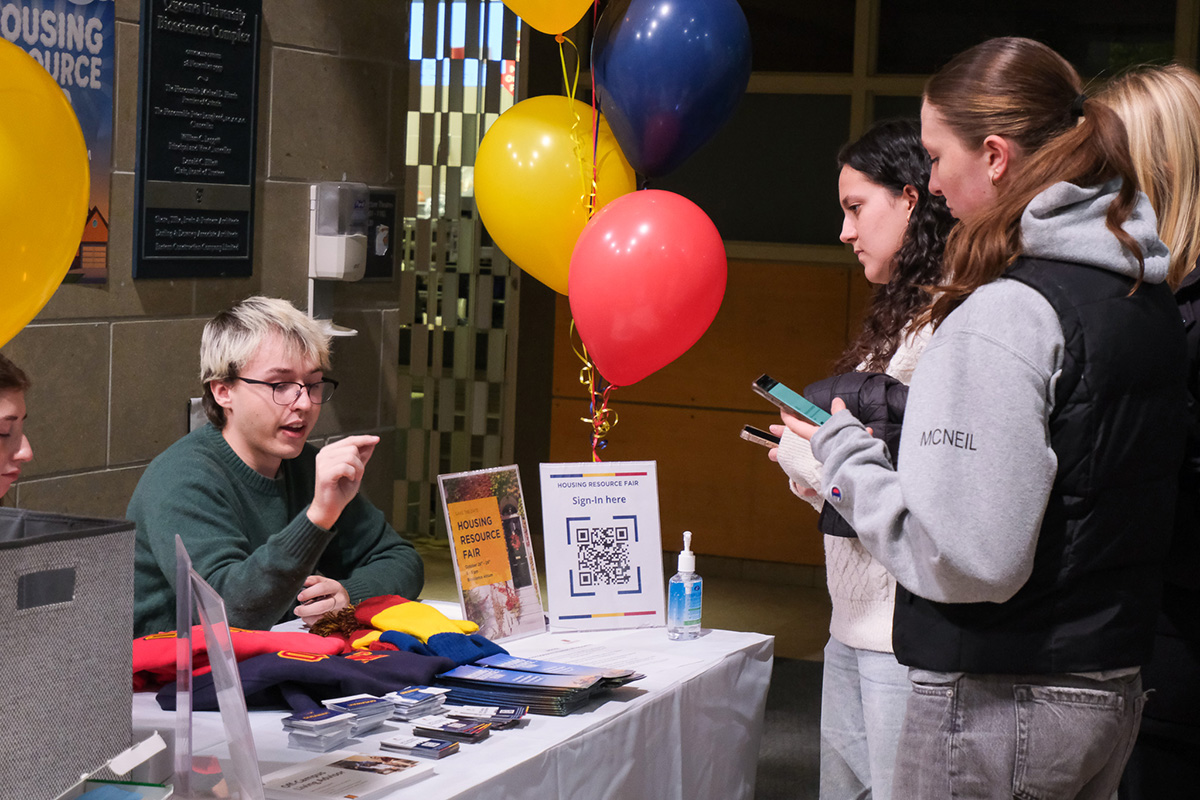 OCLA student team talking to students across a table with Queen's merchandise, pamphlets, and balloons