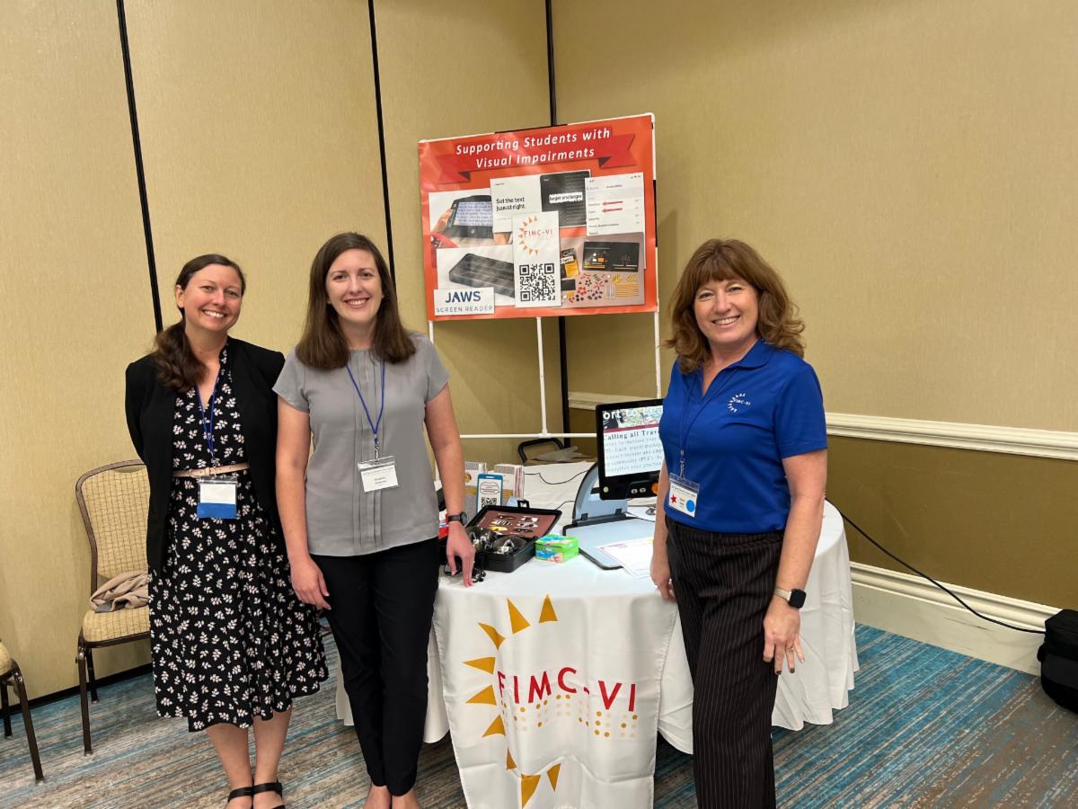 Photo of Andrea Wallace, Liz Anderson, and Cheri Hart standing in front of a table with various assistive technology devices at the SESI AT Expo.