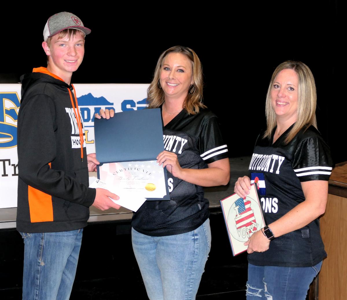 Image of Brody shaking hands with Jenny Thomas and Sarah Williams behind her. Holding his certificate for winning.