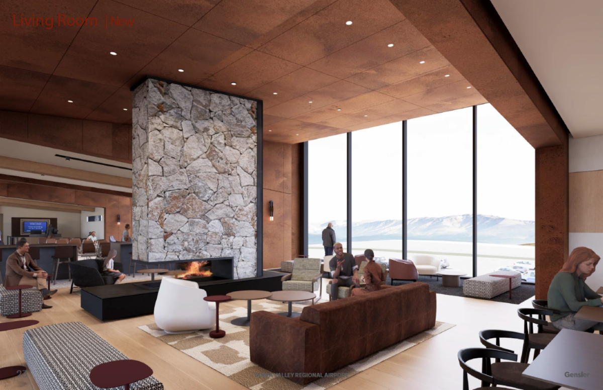 Modern living room with a large stone fireplace, high ceilings, and floor-to-ceiling windows offering views of mountains and a body of water. The space features contemporary furniture including brown leather sofas, white chairs, and patterned ottomans. Several people are seated, conversing or relaxing.