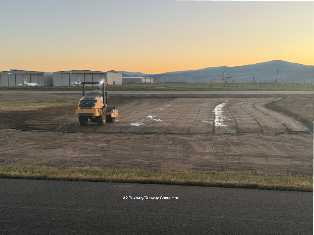 Construction scene at an airport during sunset, featuring a yellow road roller working on a prepared ground surface. Hangars and small airplanes are visible in the background. Text reads 'A2 Taxiway/Runway Connector.