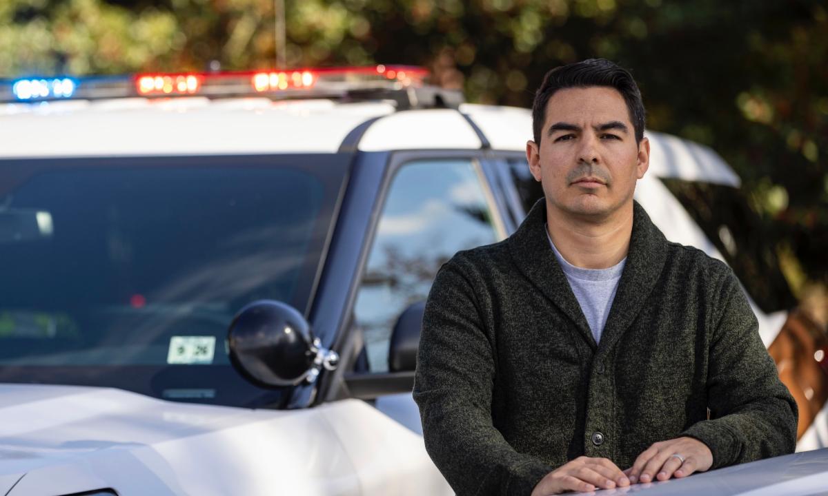 John Shjarback stands outdoors in front of a police vehicle with emergency lights visible in the background.