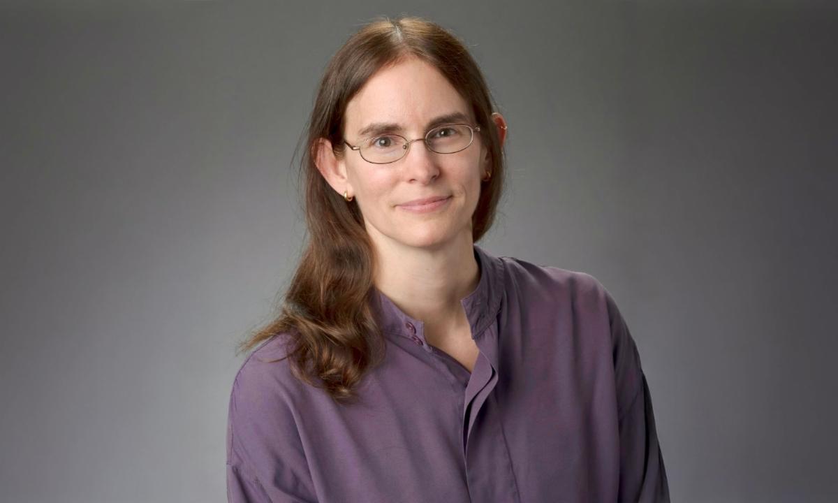 Studio portrait of Suzy Kohout wearing glasses and a long‑sleeved blouse against a gray background.