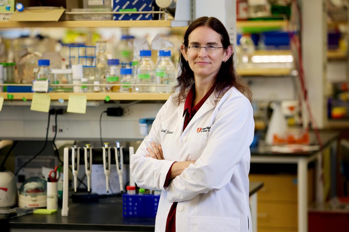 Researcher in a lab coat stands a laboratory workstation with arms crossed.