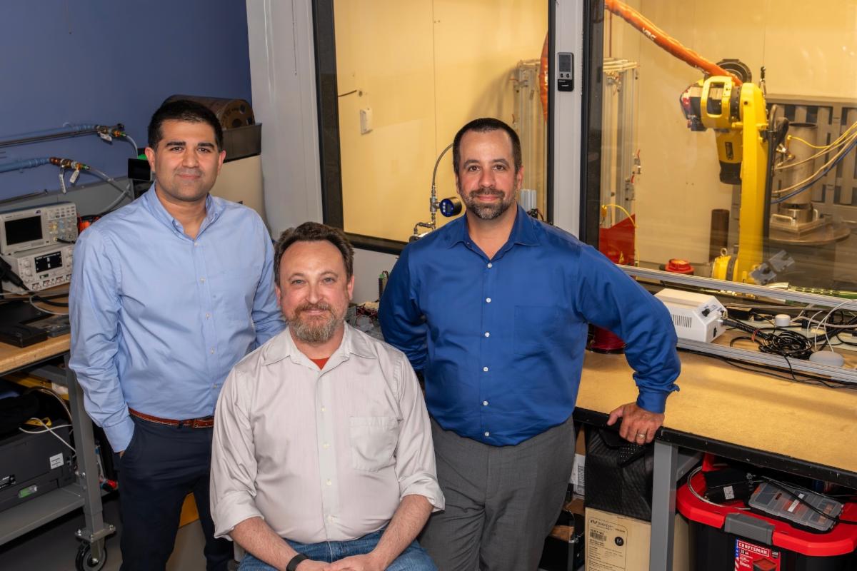 Three people in an engineering laboratory with equipment and a robotic arm in the background.