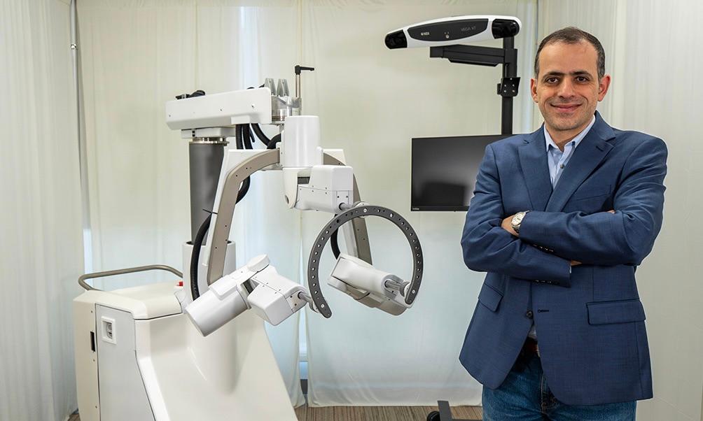 Mohammad Abedin Nasab stands in a laboratory beside Robossis Alpha, a robotic surgical system featuring articulated mechanical arms, a curved positioning component, and a mounted display screen.