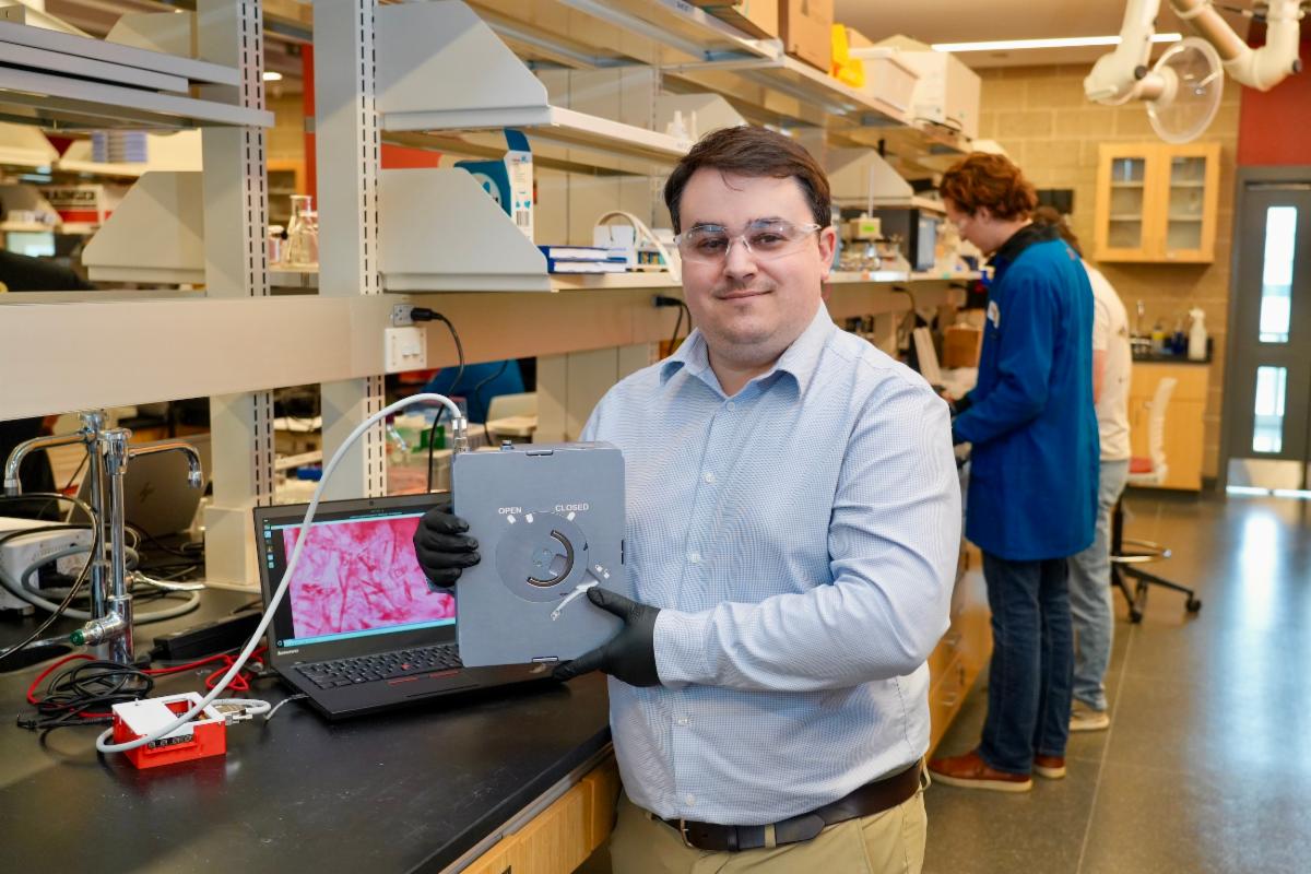 Gerard Capellades holds equipment at a bench with research equipment and colleagues in the background.