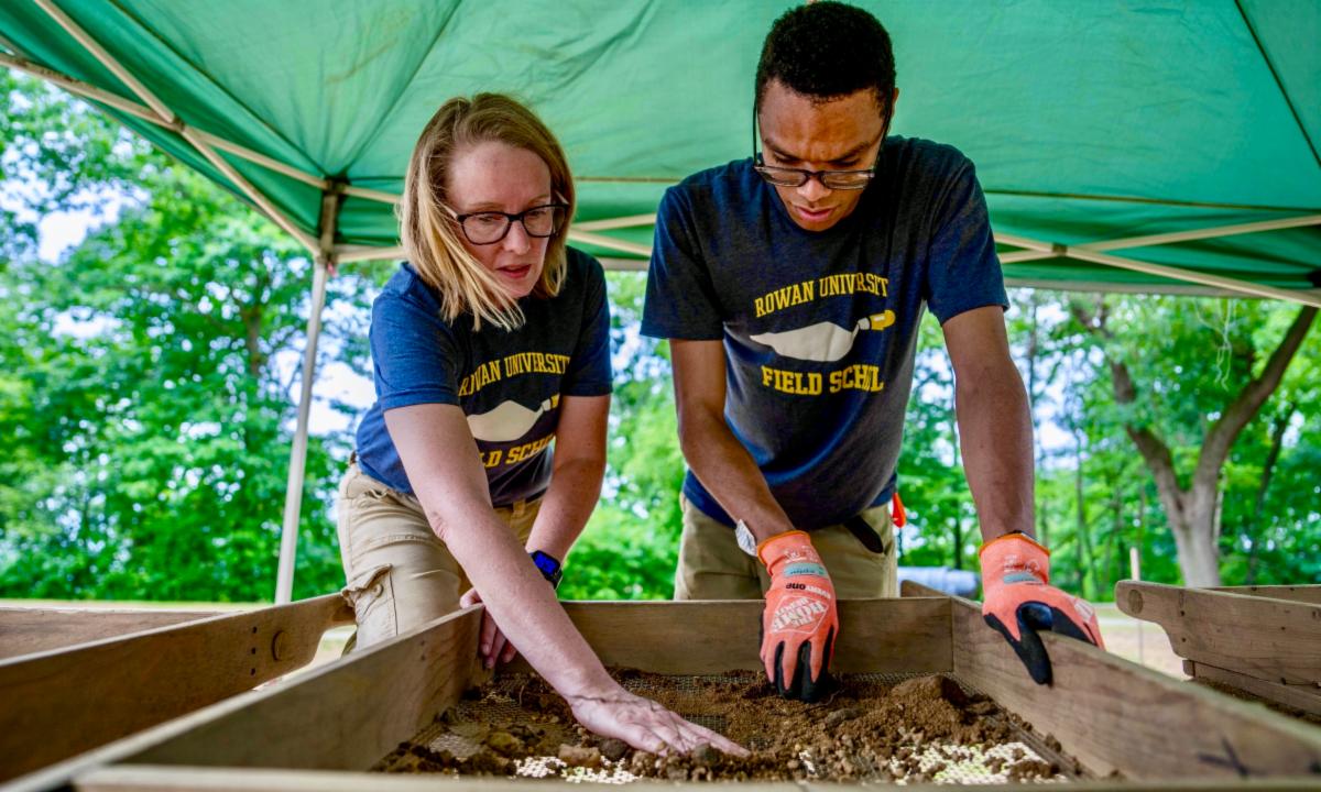 Professor and student sift soil using a wooden screen under an outdoor canopy.