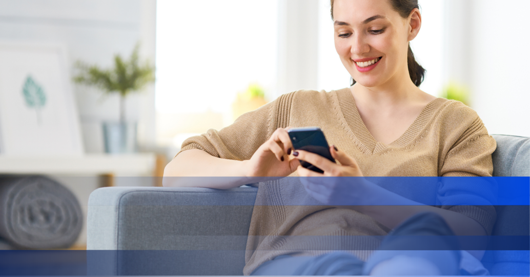 A woman sitting on a couch looking at her smart phone while smiling. 