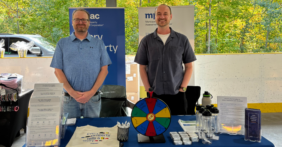 A photo of Government Relations team members standing behind the MPAC exhibitor booth at a community event, with educational materials and branded giveaways displayed on the table in front of them. 