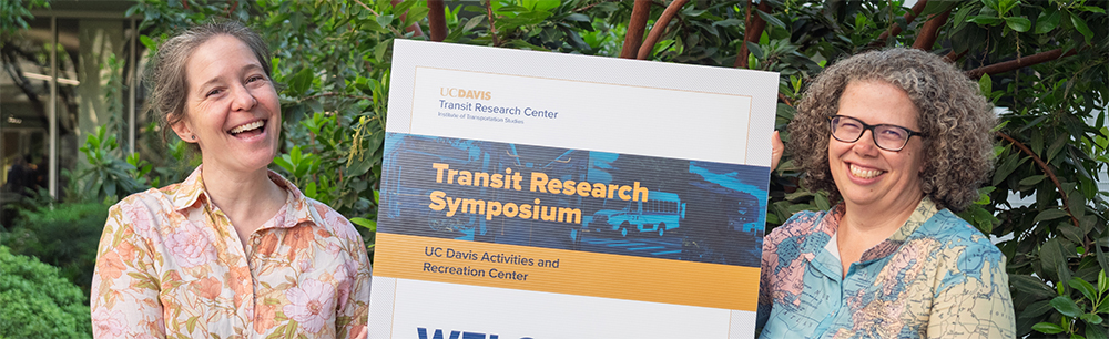 Susie Pike and Kari Watkins hold a sign for the 5th annual Transit Research Symposium.