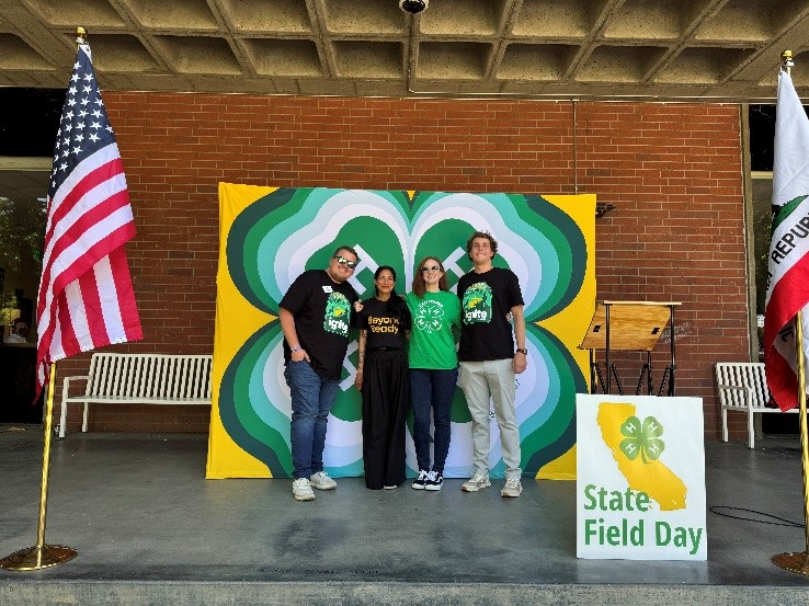 Members of the Events and Youth Experiences team from National 4-H Council pose on front of a brick wall and large 4-H Beyond Ready banner. The U.S. flag is on one side, the California flag is on the other side and there is a California 4-H State Field Day sign.