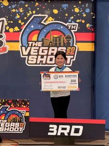 4-H youth member standing on the event podium holding a 3rd place scholarship award at the Vegas Shoot 2026 archery competition. The youth is standing in front of the event logo, which shows the the Las Vegas skyline and "60th". The backdrop is decorated with red and yellow stripes, and blue, yellow and red splatter design.