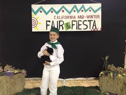 Young Addison Holbrook holding a rabbit that she showed at the mid-winter Fair in Imperial County. Addison is wearing white pants, white shirt, the California 4-H green hat and scarf around her neck and shoulders.