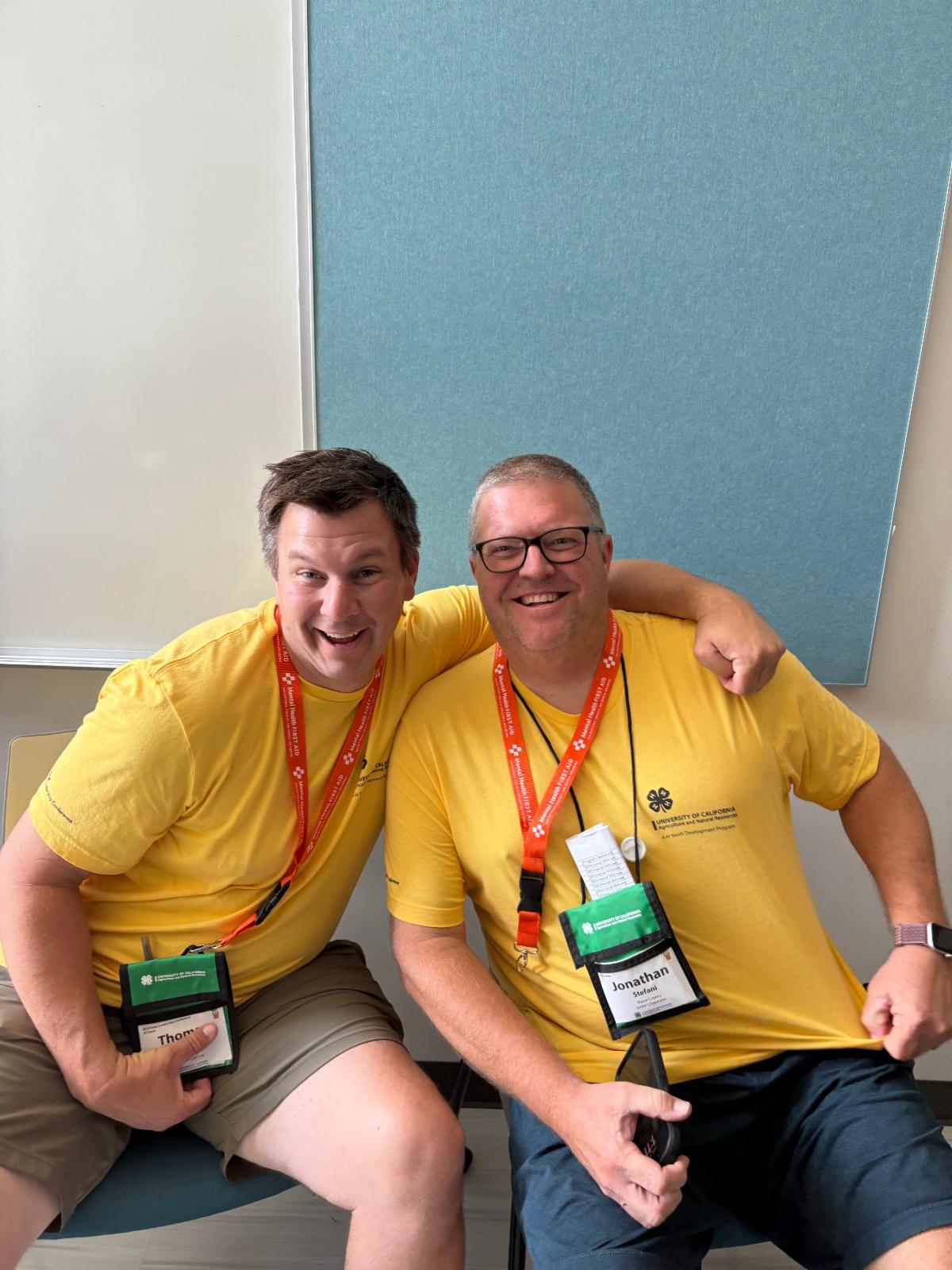 image depicts 2 4-H adults sitting on chairs, posing for the photo. Theperson on the left has their left arm around the shoulders of the person on the right. Both are wearing SLC shirts, lanyards and name badges. There is a blue bulletin board on a white wall behind them.