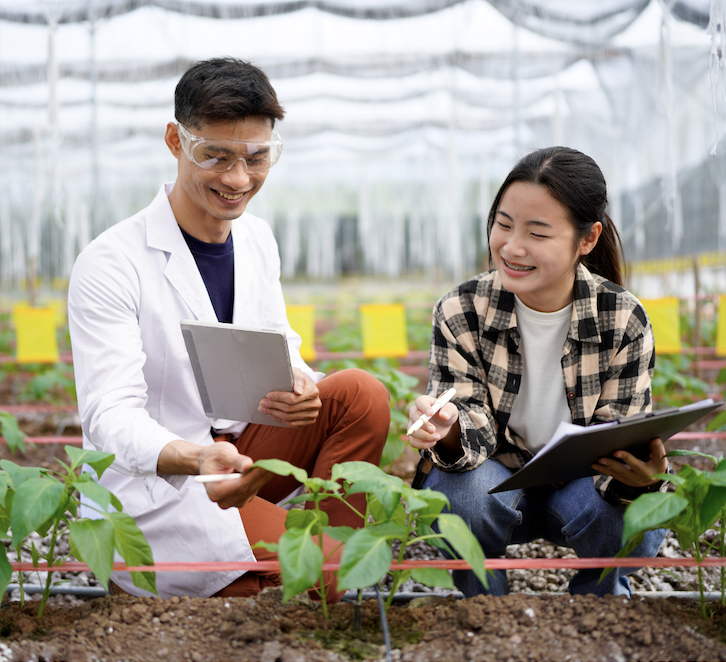 Image shows two people in a greenhouse, crouching in front of small sprouting plants. One person is wearing safety glasses and a white lab coat, showing a feature of a leaf to the other person. The other person is dressed in casual clothing, holding a pen and clipboard. Behind them are several rows of plants, and above is the structure of the greenhouse.