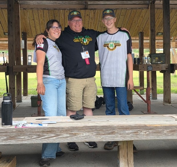 Modoc County Shooting Sports volunteer Bill Engle standing under a wooden structure with two other individuals in a smiling pose. All three people are wearing shirts associated with the California 4-H Shooting Sports program.