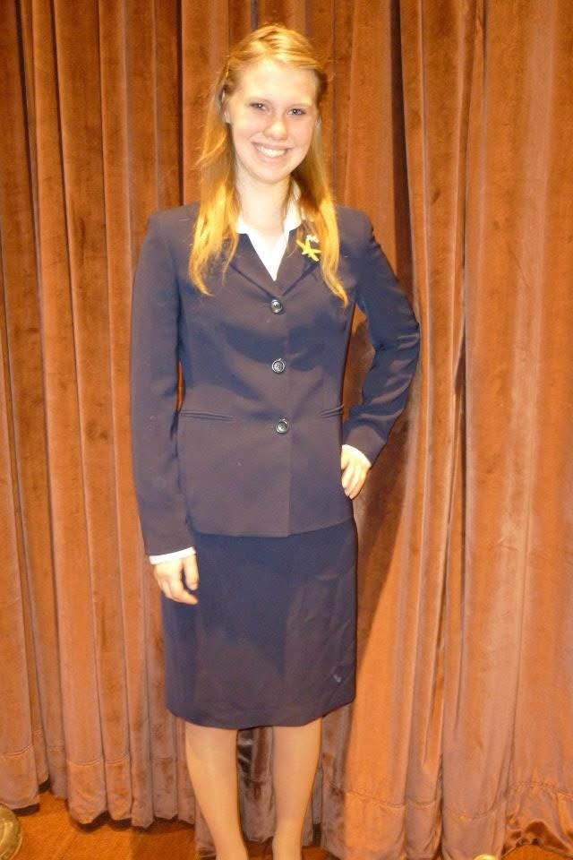 Kate Lyn Sutherland as a State Ambassador standing wearing a blue shirt and suit jacket.