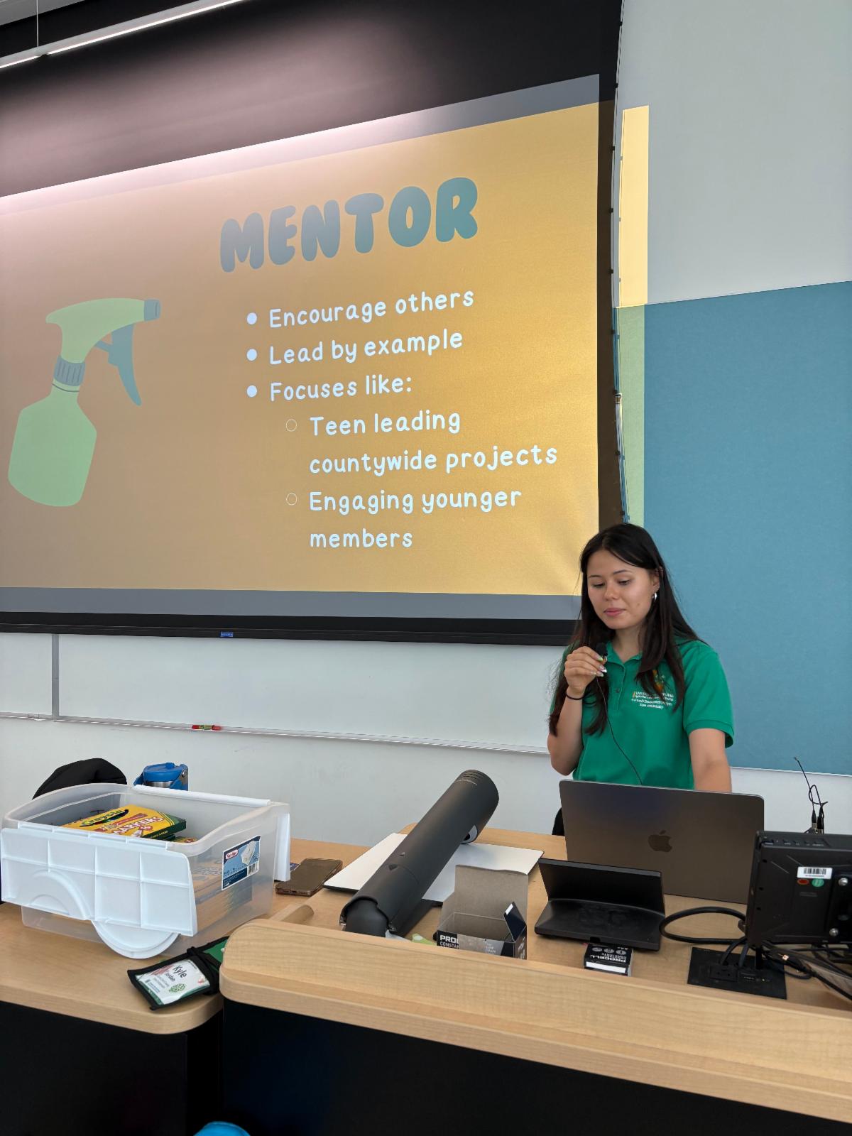 Image shows a 4-H state ambassador standing in front of a desk with an open laptop. There is a plastic container with art supplies on an adjacent table, and a drop-down projector behind her. On the projector is an image that reads Mentor: encourage others, lead by example, focuses like teen leading countywide projects, engaging younger members.