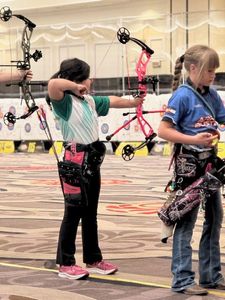 4-H youth members compete at an indoor archery contest. One youth is turning away carrying archery equipment. The other youth is aiming a bow. In the distance is a wall of targets of varying size and color.